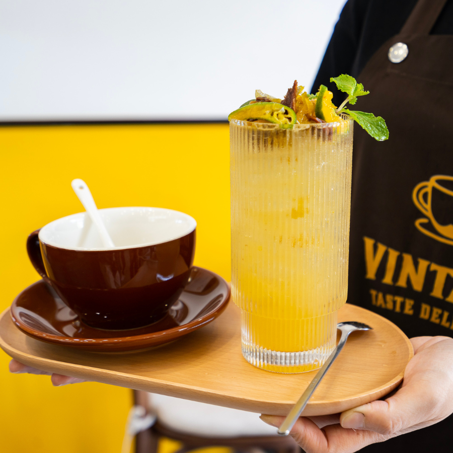 Nhân viên phục vụ tại Vintage Taste Deli Cafe cầm khay gỗ với ly nước vàng tươi và tách cafe nâu sang trọng.
